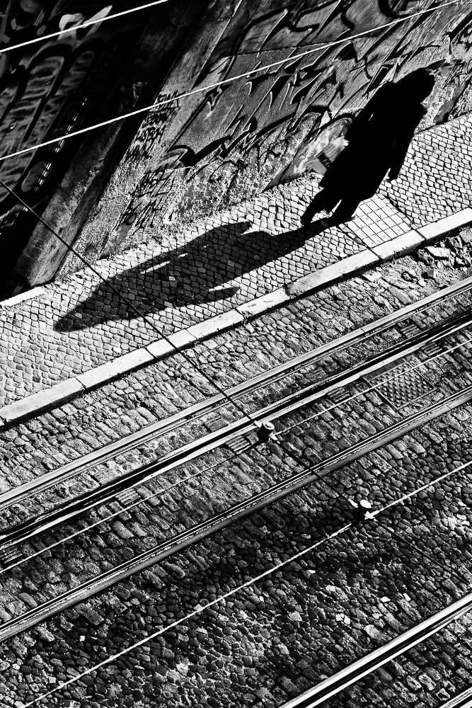 Fot en blanco y negro y desde arriba de la subida a la Gloria, en Lisboa. Se ve una persona en la acera, proyectando su sombra paralela a los railes del tranvía.