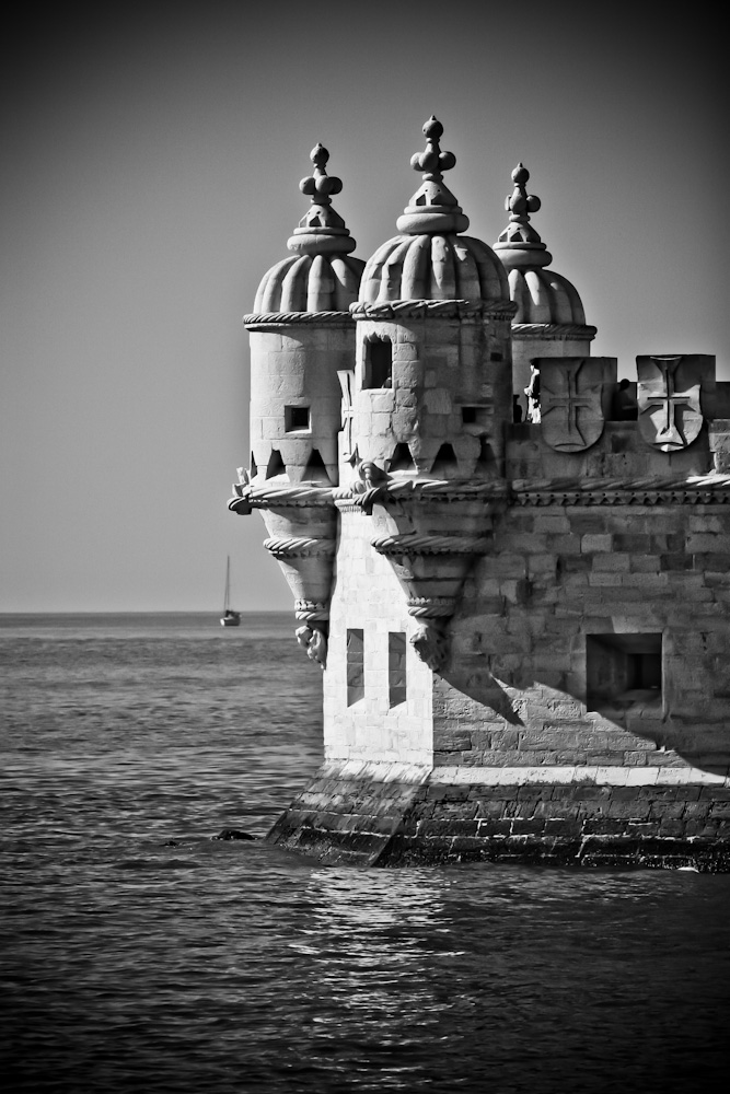 Fotografía en blanco y negro de parte de la Torre de Belem sobre el agua y mirando a mar abierto por donde asoma un velero