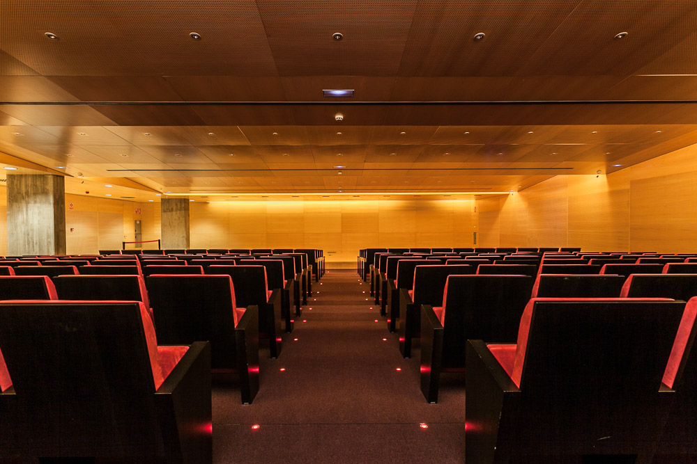 Fotografía de salón de actos con techos y paredes de madera y sillones tapizados en rojo